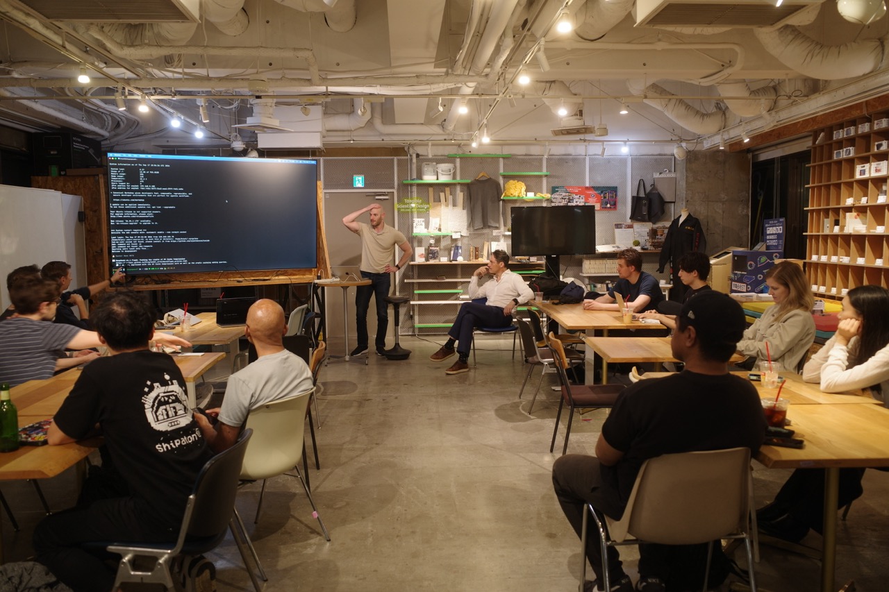 A presenter standing beside a terminal on the big screen while attendees watch from the tables at FabCafe